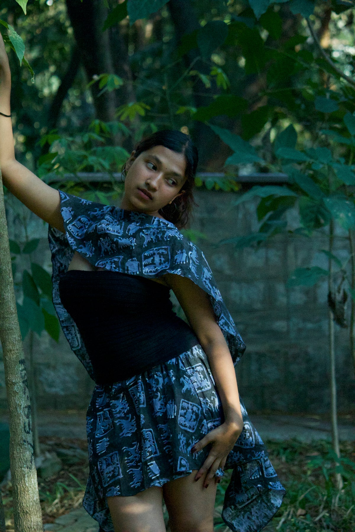 Woman in a patterned dress standing in a forested area with a wall in the background
