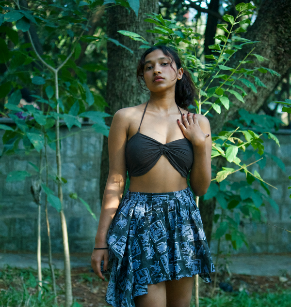 A woman stands outdoors in a wooded area wearing a black halter crop top with a twisted front detail and a high-waisted, asymmetrical blue and black patterned skirt.