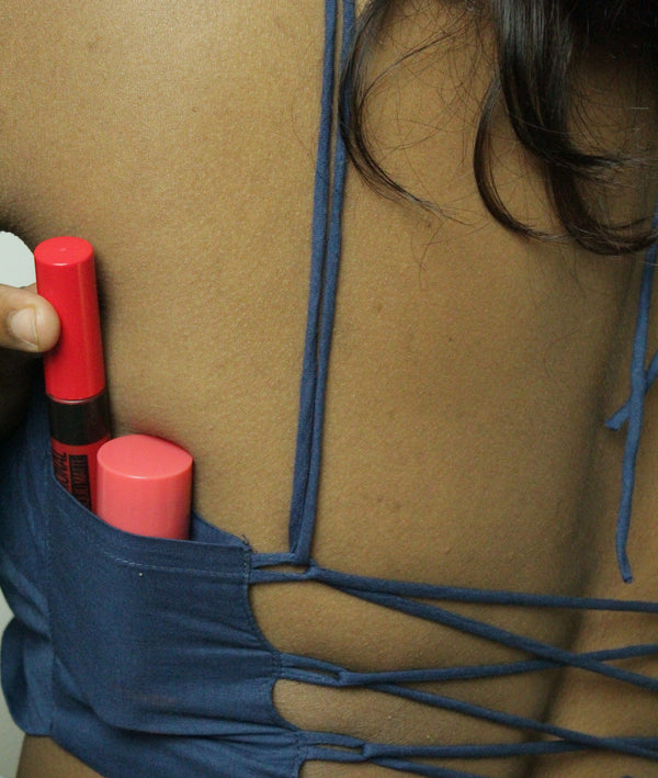 Close-up of a person's bare back wearing a dark blue strappy top, with a hand holding two red cosmetic tubes in the side pocket.