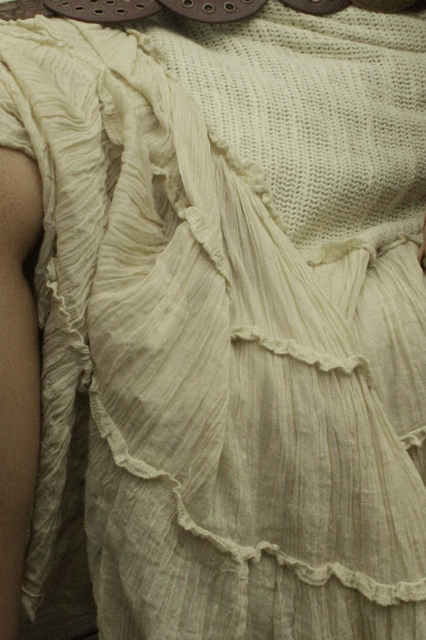 A person seated in a wicker chair wearing a cream or off-white tiered skirt with a large brown belt featuring round metallic medallions.