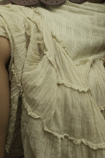 A person seated in a wicker chair wearing a cream or off-white tiered skirt with a large brown belt featuring round metallic medallions.