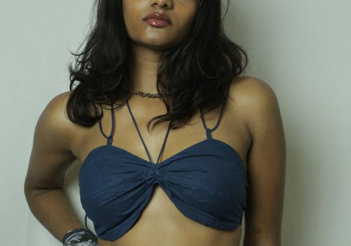 A woman modeling a dark blue twisted-front bandeau-style top with thin straps, paired with a gray patterned skirt, standing against a plain light wall.