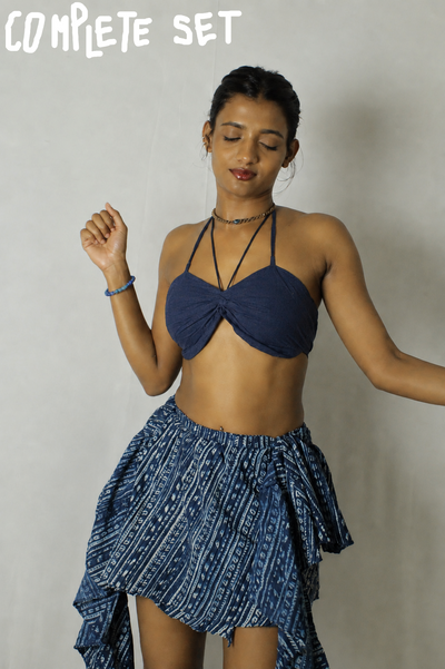 A woman modeling a two-piece blue and white patterned set consisting of a halter crop top and a matching high-low ruffled skirt, set against a plain light gray wall.