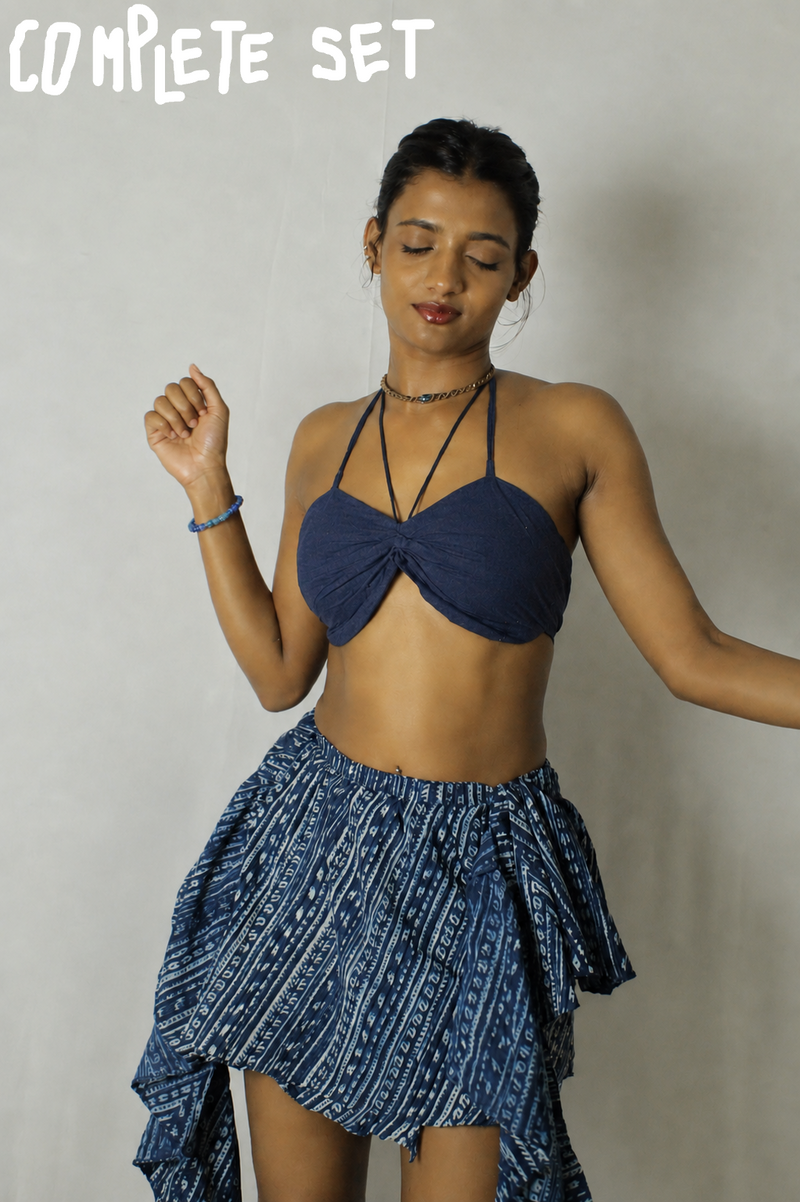 A woman modeling a two-piece blue and white patterned set consisting of a halter crop top and a matching high-low ruffled skirt, set against a plain light gray wall.