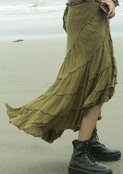 A person standing on a beach wearing a long, tiered olive green skirt, a brown patterned top, and black lace-up boots.