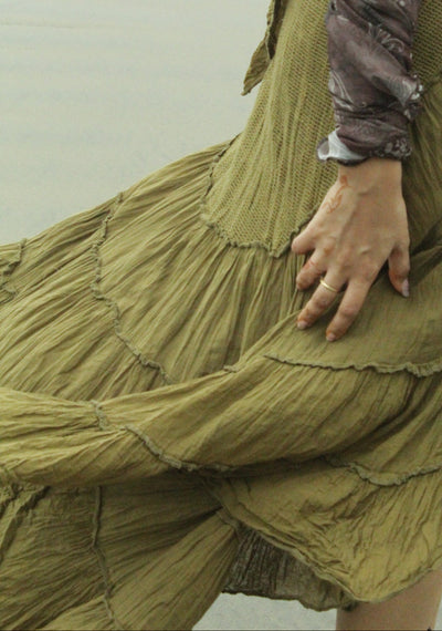 A close-up of a person outdoors wearing a long, tiered olive green skirt and a patterned brown top, with one hand resting on the skirt.
