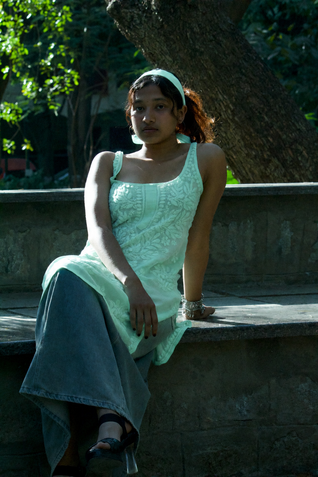 A woman sits on a stone ledge outdoors, wearing a mint green sleeveless embroidered dress with a matching headband and a long denim skirt.
