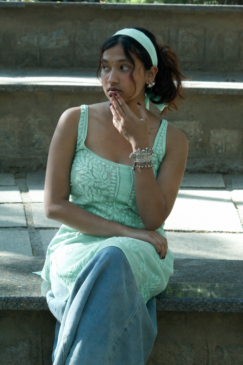 A woman sitting on outdoor stone steps wearing a mint green embroidered sleeveless top and matching headband, with blue jeans and silver bracelets.