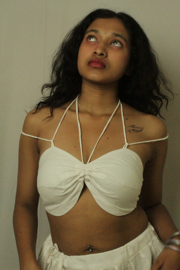 A woman wearing a white halter crop top with a front twist detail and matching white bottoms, standing against a plain light gray wall.