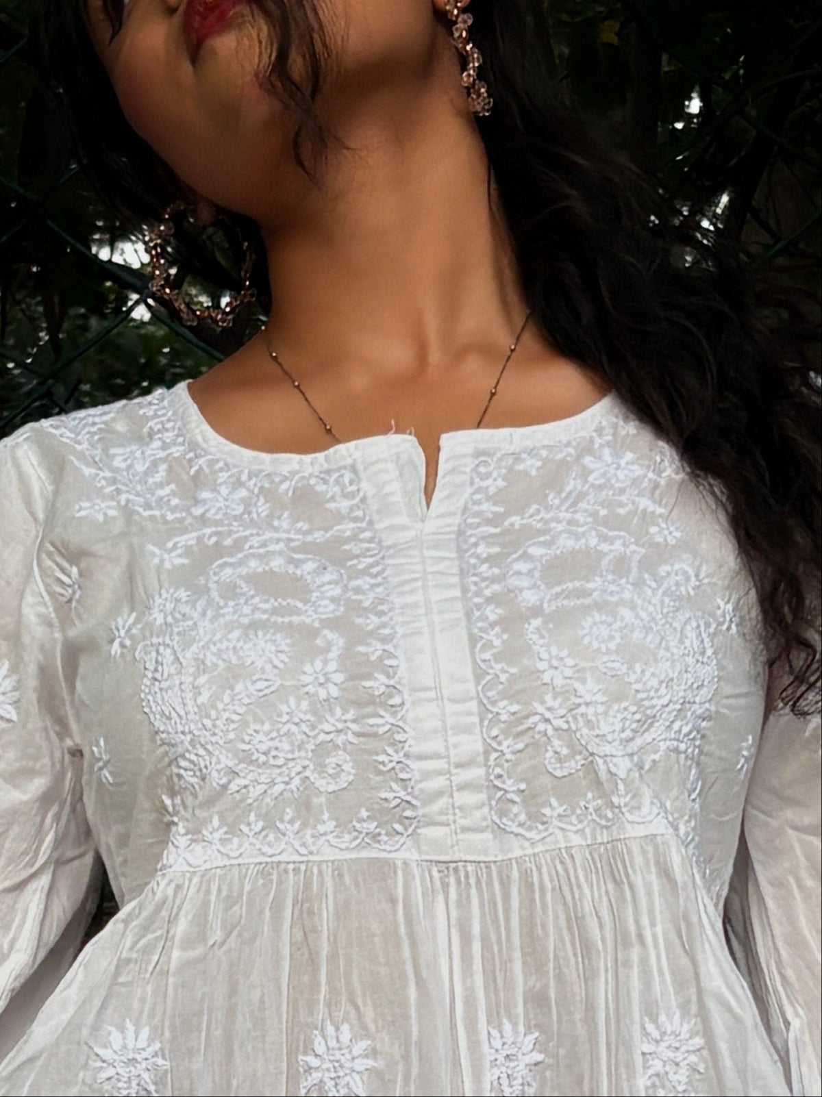 A close-up of a woman wearing a white long-sleeve embroidered dress with intricate floral lace detailing on the bodice and neckline.