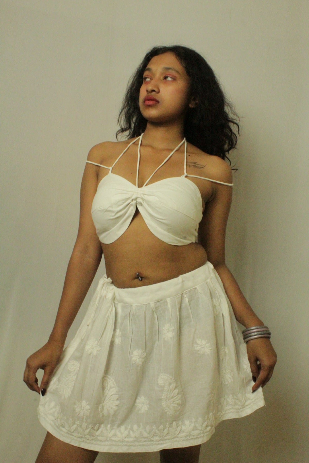 A woman modeling a white two-piece outfit consisting of a halter crop top with a twisted front detail and a matching embroidered mini skirt, set against a plain light gray wall.